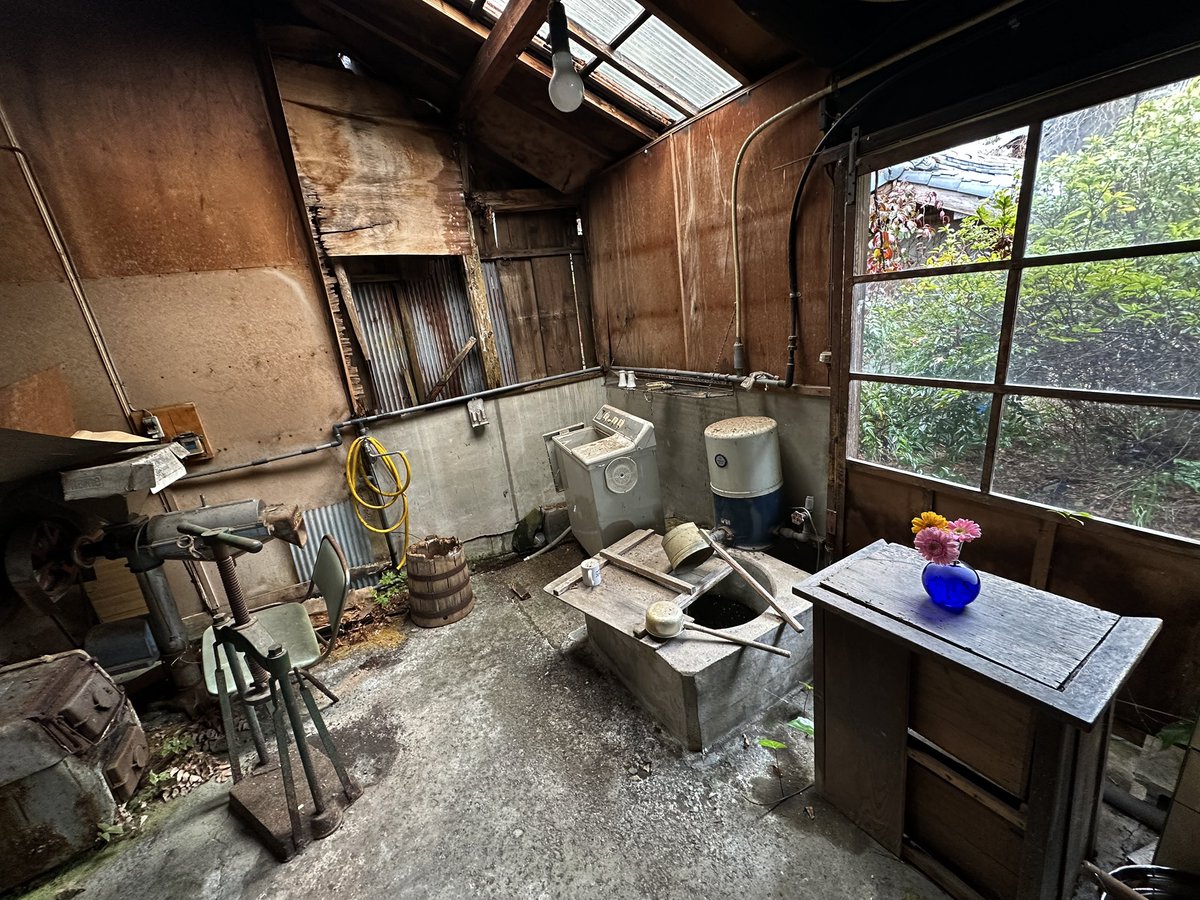 古民家内部の作業場風空間に青い花瓶が置かれ、錆びた道具や窓からの緑と融合。生活の痕跡が残る会場でアートが日常に溶け込む様子を捉えた写真