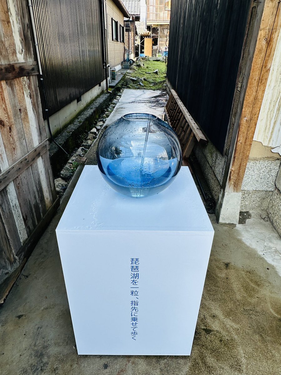 路地に設置された白い台座上の大型青いガラス球体、水滴がつき側面に「琵琶湖を一粒、指先に乗せて歩く」との文字。古民家と水のインスタレーションが融合した新作風景