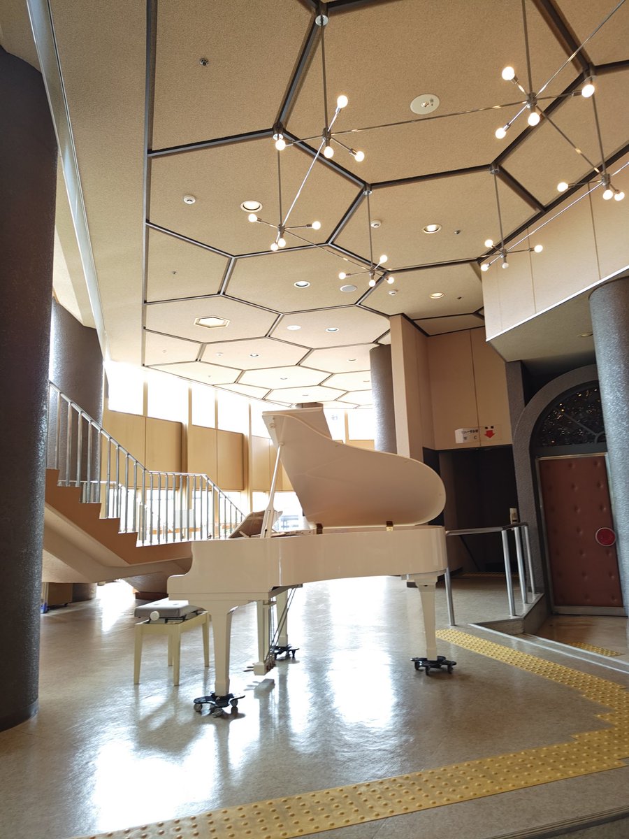 高島市民会館ロビーに設置された白いグランドピアノ周辺の様子。吹き抜けのロビーに自然光が入る開放的な空間で、誰でも演奏できるストリートピアノ企画の雰囲気を伝える写真。