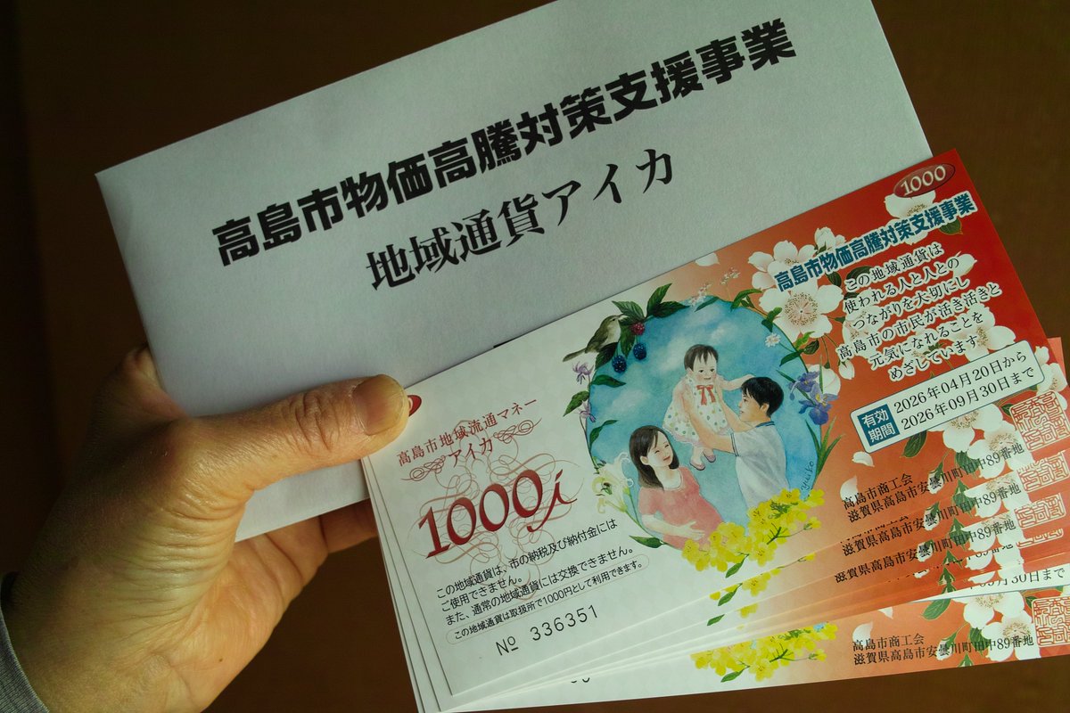 高島市物価高騰対策支援事業として配付された地域通貨「アイカ」の実際の現物写真。白い封筒に「高島市物価高騰対策支援事業 地域通貨アイカ」と記載され、手に持った1,000円券（家族イラスト入り、額面1000i、有効期間2026年4月20日～9月30日）が鮮明に写っている。市民が受領した直後の様子を捉えた実物ビジュアル。
