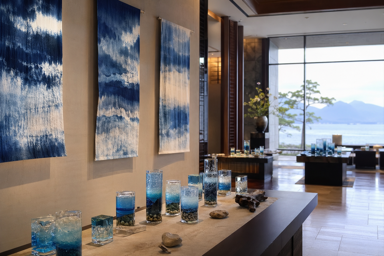 滋賀県高島市今津サンブリッジホテル1階ロビーギャラリーで開催された中條弘之個展「水辺の贈り物」の会場風景。琵琶湖の砂や小石を封じ込めたジェルキャンドルと、水の流れを表現した染織作品が並ぶ展示空間を正面から捉えたもの。柔らかな光が作品に反射し、湖の静けさを思わせる雰囲気。