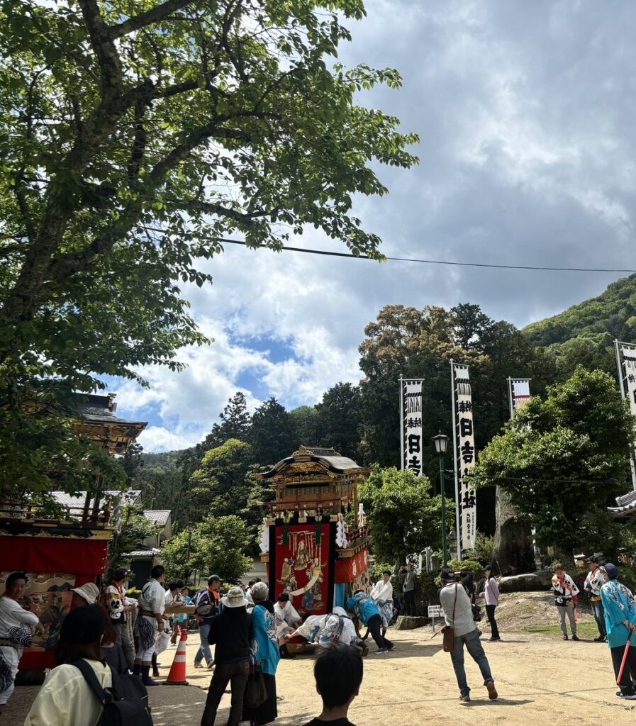 日吉神社周辺で曳山と旗が並ぶ大溝祭の賑わい。青空の下、木々と山々を背景に伝統行事が展開される様子で、湖西の自然と文化の調和を示す。